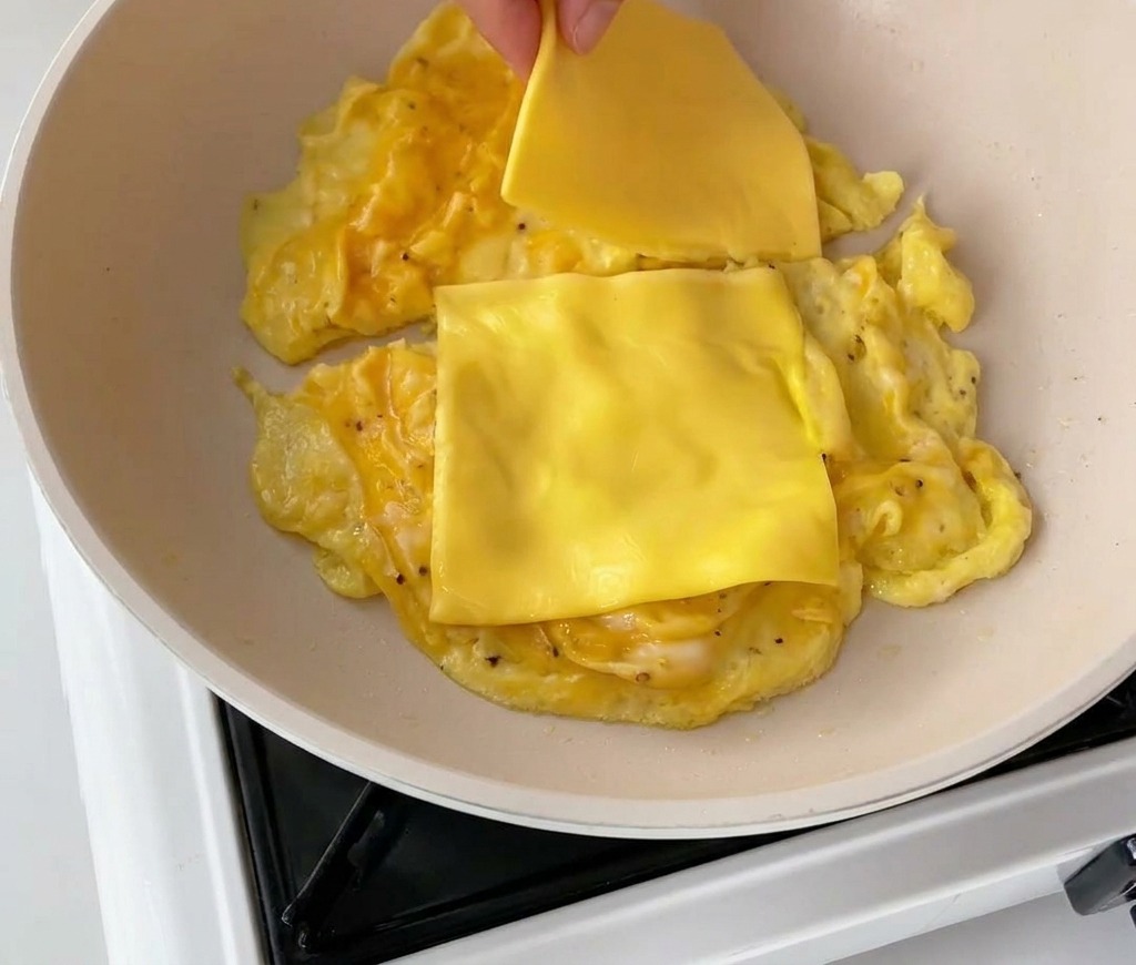 Une main déposant deux tranches de cheddar sur les œufs brouillés dans la poêle.