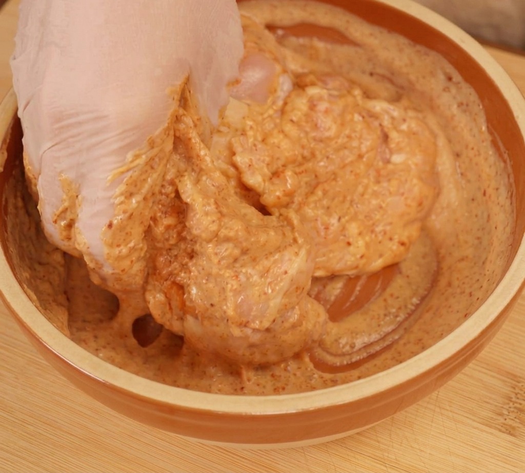 Une main gantée mélangeant soigneusement le poulet dans une marinade crémeuse orangée.