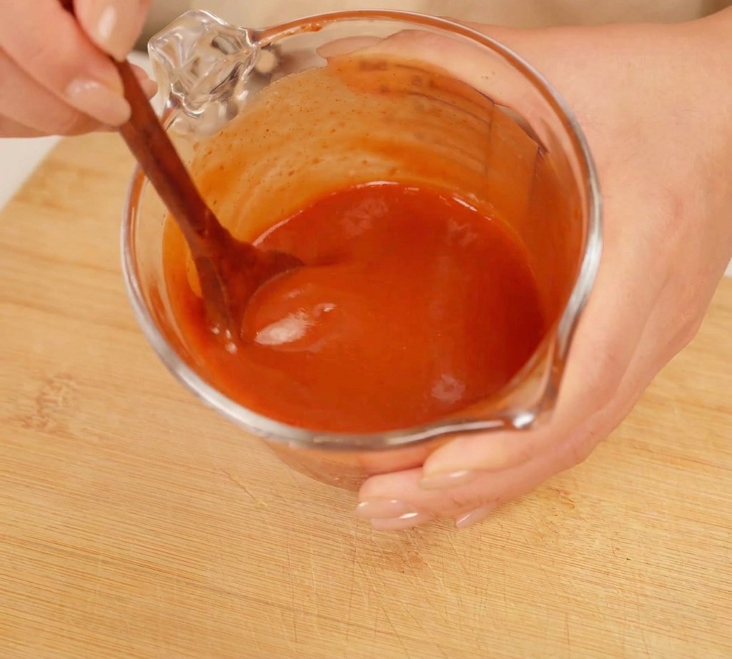 Une cuillère en bois mélangeant une sauce rouge épaisse dans un verre doseur.