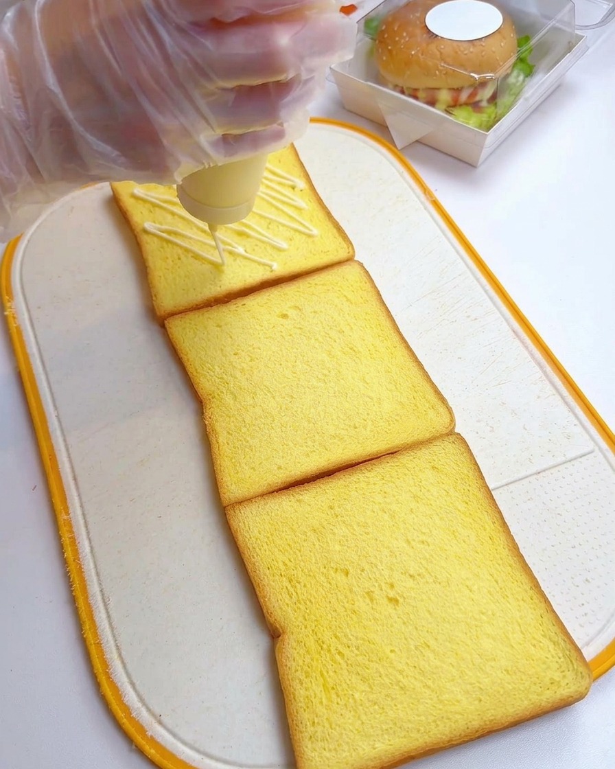 Main gantée versant de la mayonnaise en zigzag sur trois tranches de pain de mie.