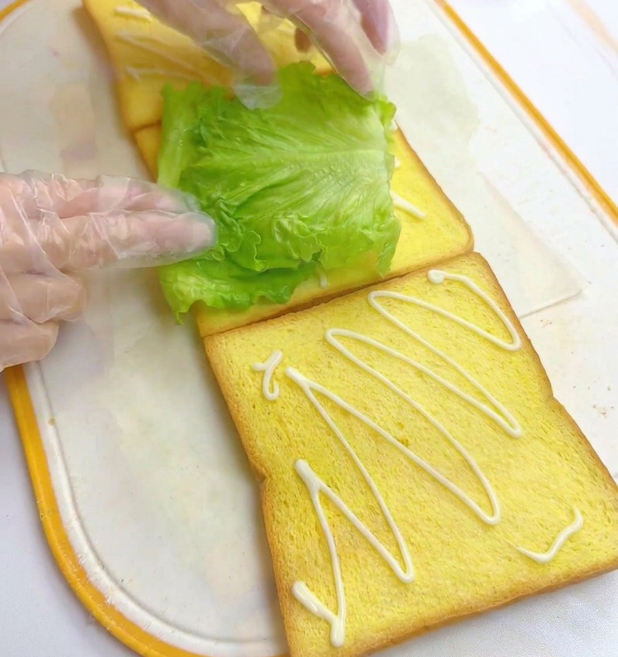 Mains plaçant une feuille de laitue fraîche sur une tranche de pain nappée de mayonnaise.