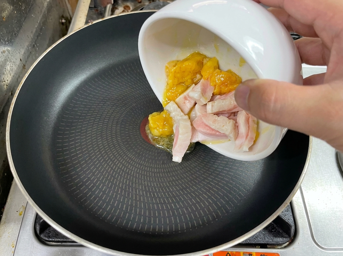 Main versant des morceaux de gras de poulet et de porc dans une poêle noire.