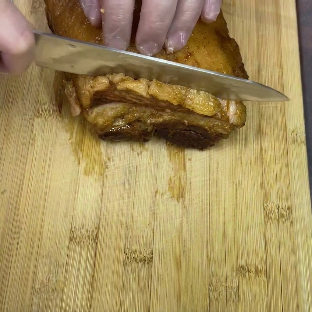 Mains gantées coupant une pièce de poitrine de porc braisée sur une planche en bois.