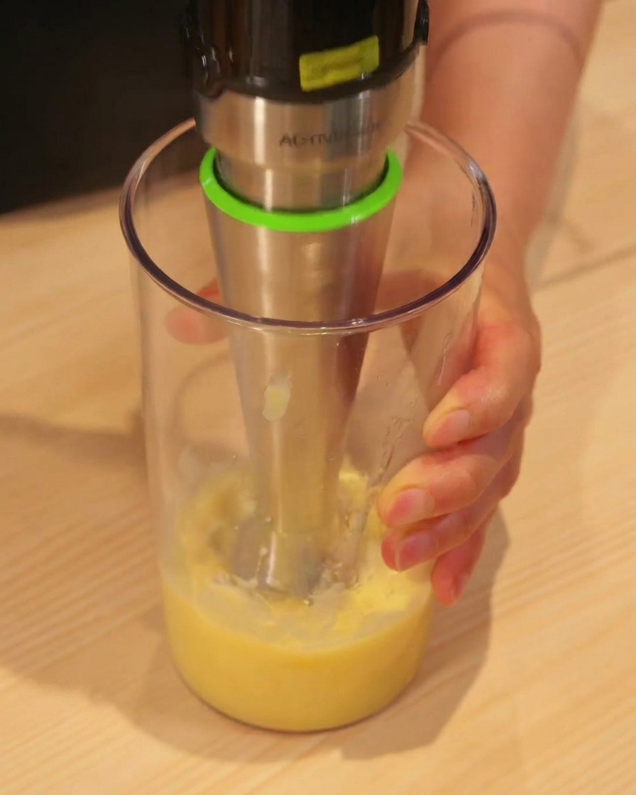 Un mixeur plongeant transformant des morceaux dananas jaune en jus lisse dans un grand récipient.