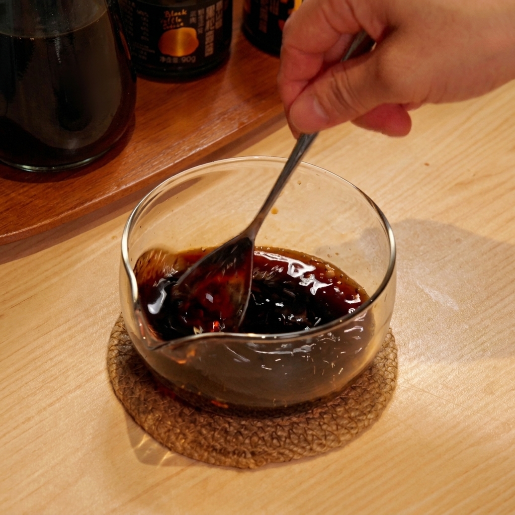 Une main utilisant une cuillère pour mélanger une sauce sombre dans un petit bol en verre.