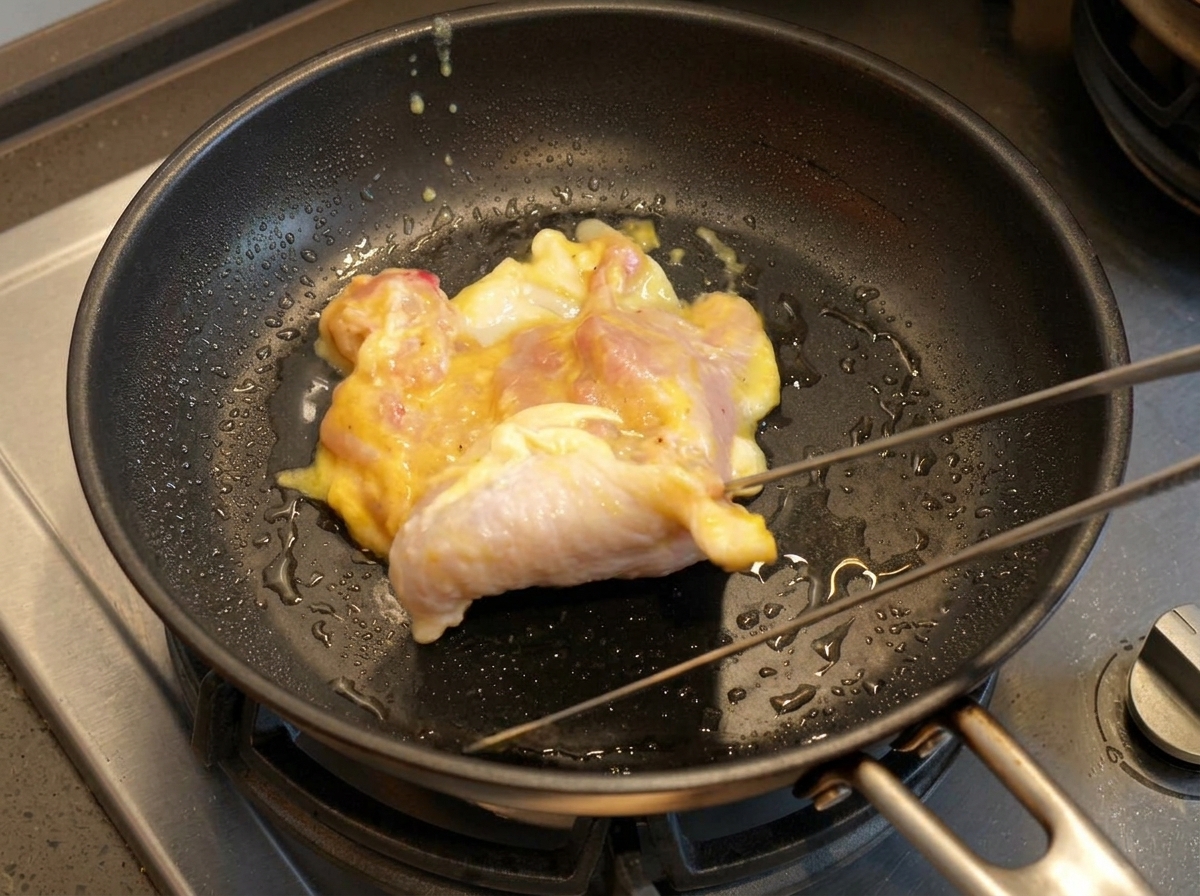 Une cuisse de poulet marinée déposée dans une poêle chaude et huilée avec des pinces métalliques.