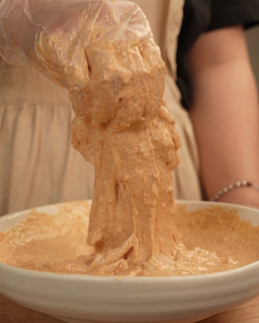 Main gantée soulevant un morceau de poulet enrobé dune pâte épaisse et collante orange pâle.