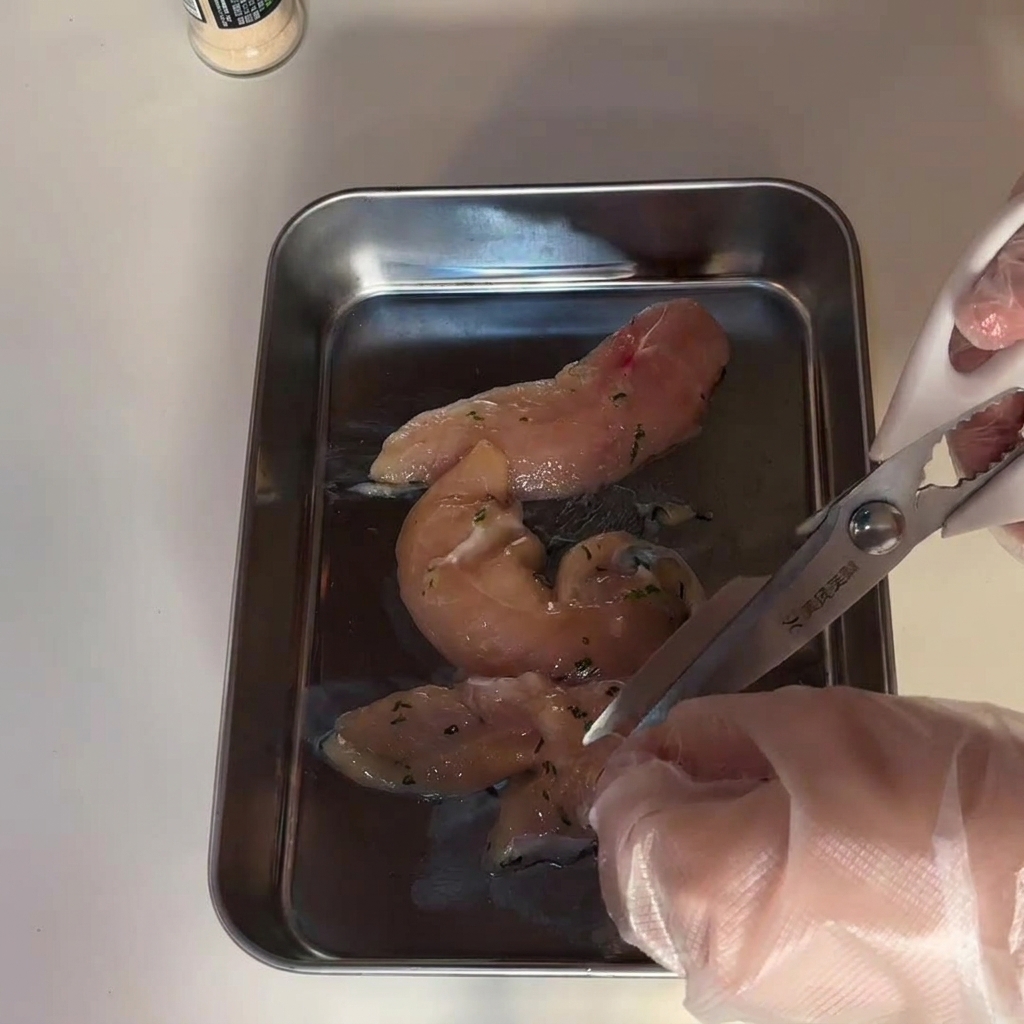 Mains gantées coupant des morceaux de blanc de poulet avec des ciseaux de cuisine sur un plateau en inox.