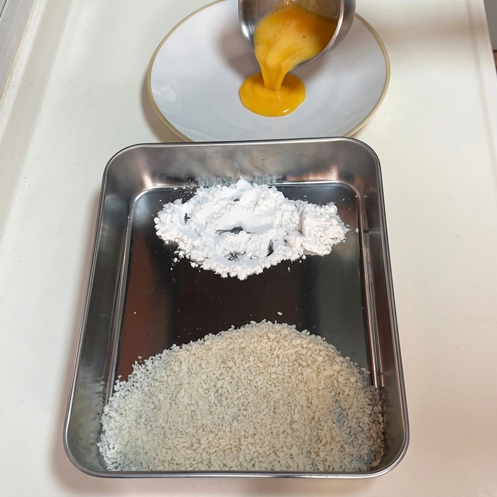 Versement dœufs battus dans une assiette blanche à côté dun plateau avec de la fécule de maïs et de la chapelure panko.