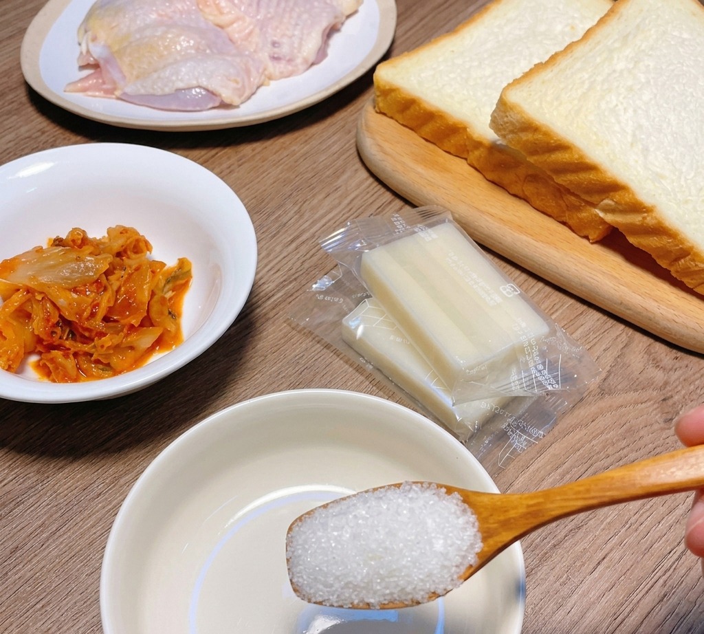Une cuillère en bois versant du sucre dans un petit bol blanc, entouré des ingrédients de la recette comme le pain et le poulet.