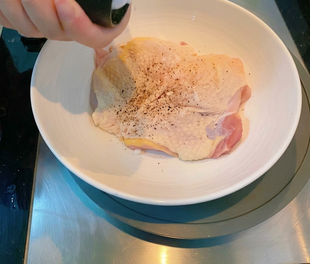 Une main utilisant un moulin à poivre pour assaisonner un haut de cuisse de poulet cru dans un bol en céramique.