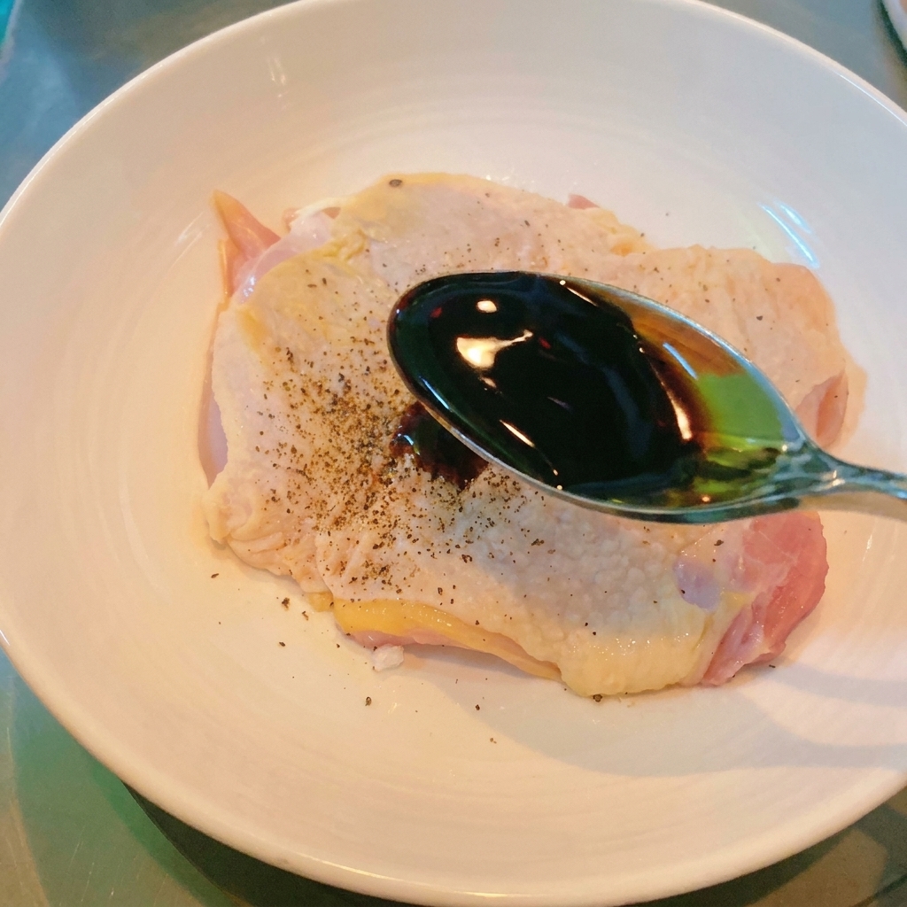 Une cuillère en métal versant de la sauce soja sombre sur le morceau de poulet assaisonné dans un bol blanc.