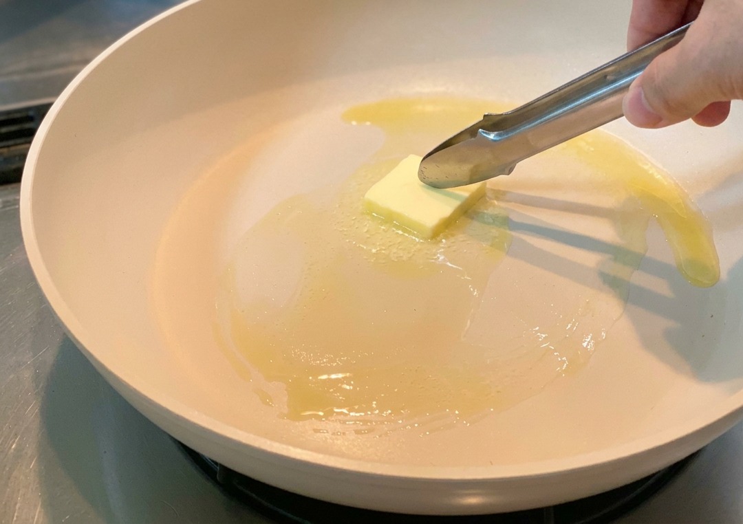 Un cube de beurre jaune fondant dans une poêle claire, manipulé avec des pinces de cuisine.