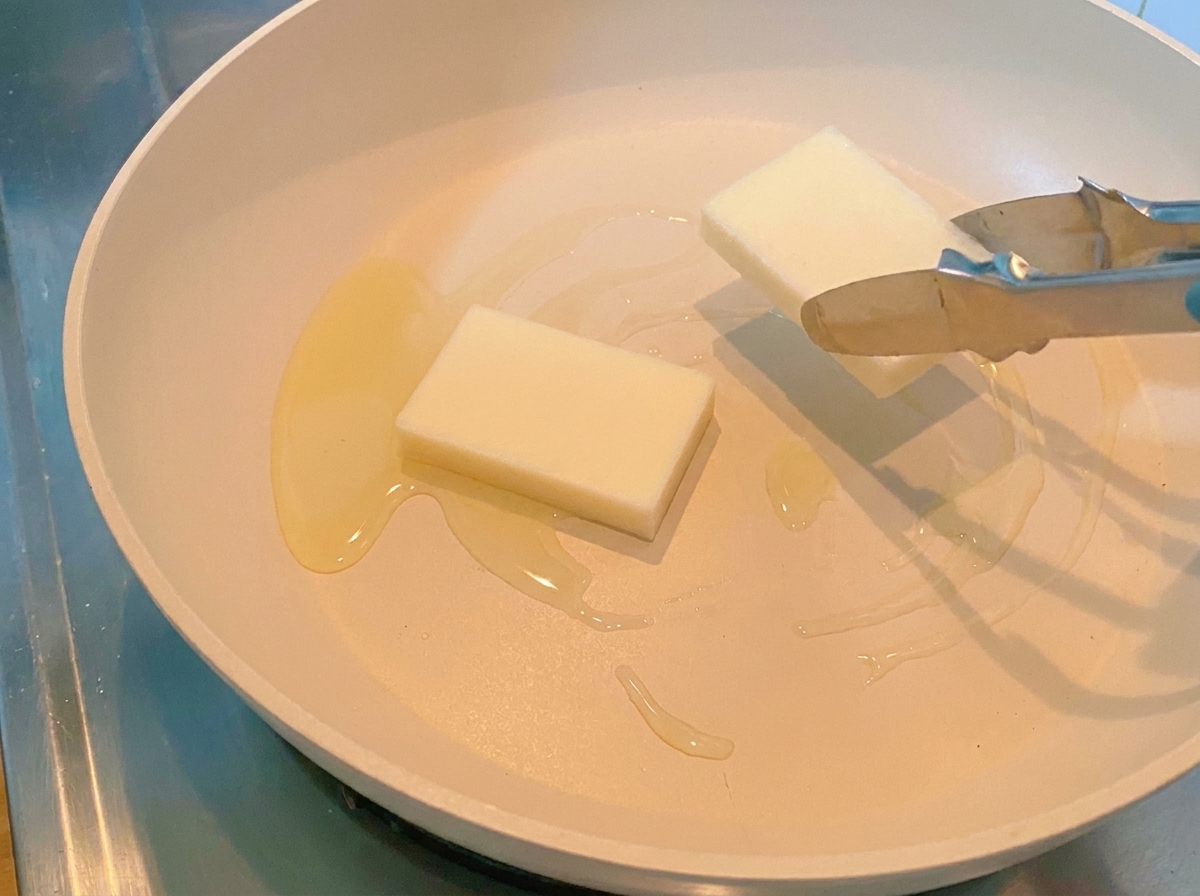 Des pinces déposant deux gâteaux de riz blancs épais et rectangulaires dans une poêle chaude.