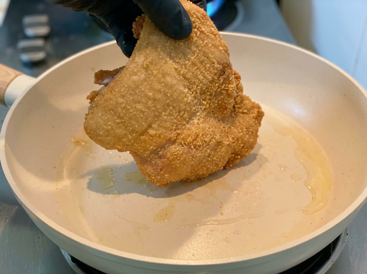 Une main gantée déposant le haut de cuisse de poulet enrobé de fécule dans la poêle chaude.