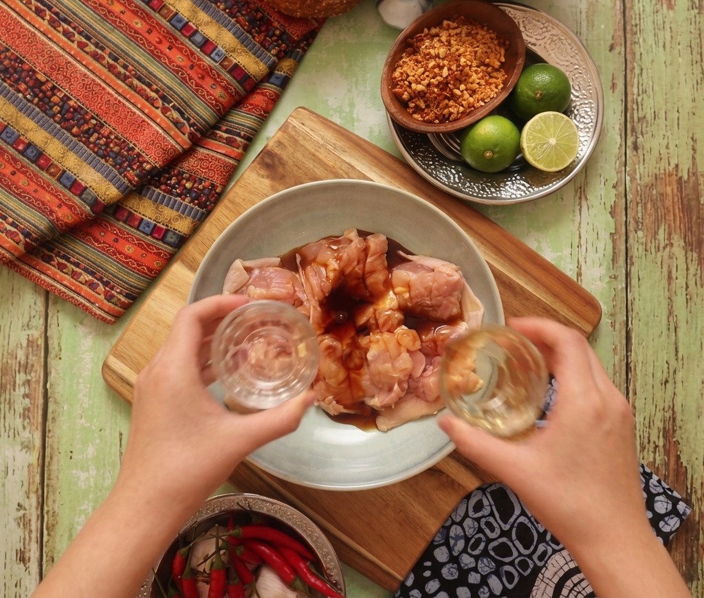 Deux mains versant une marinade liquide depuis des petits verres sur du poulet cru dans un bol bleu clair.