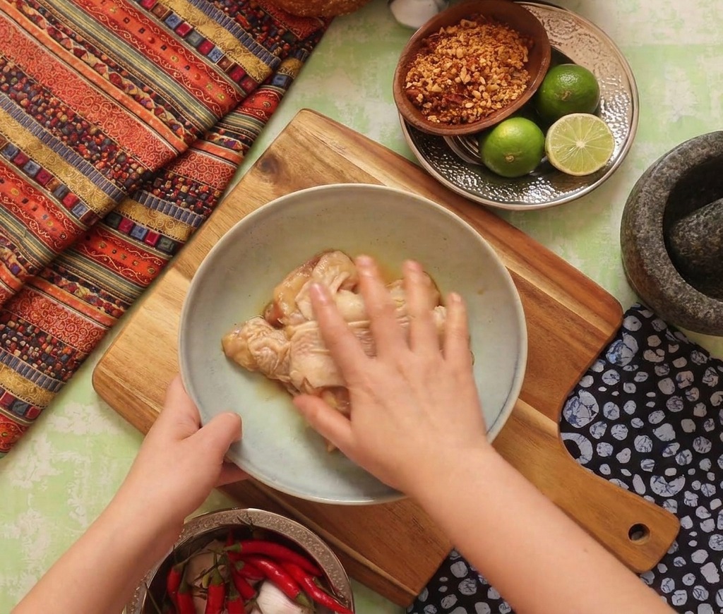 Une main massant la marinade sombre sur les cuisses de poulet crues dans un bol sur une planche en bois.