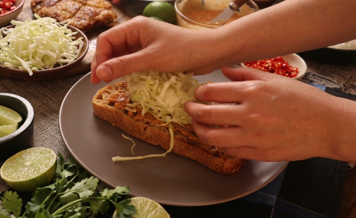 Mains déposant un monticule de chou blanc émincé sur une tranche de pain nappée de sauce.