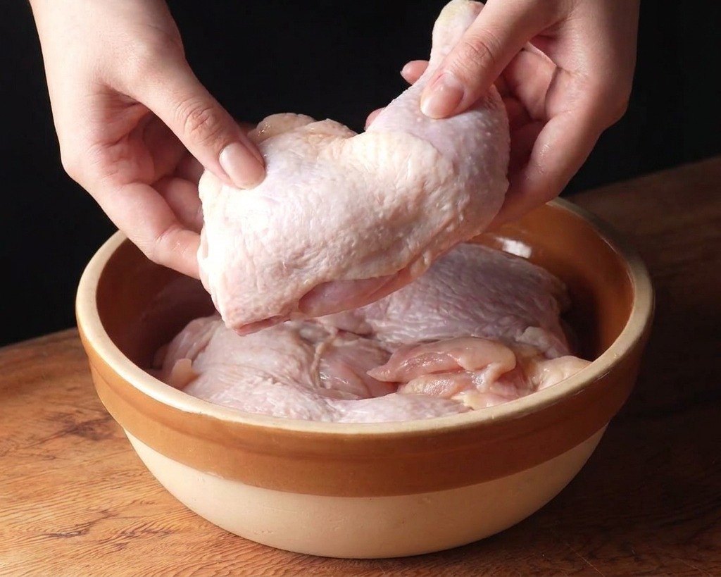 Mains plaçant des hauts de cuisses de poulet crus dans un grand bol en céramique beige.