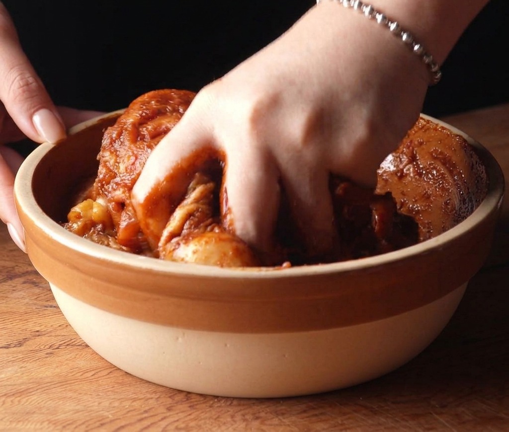 Une main mélangeant le poulet enrobé dune marinade rouge foncée dans un bol en céramique.