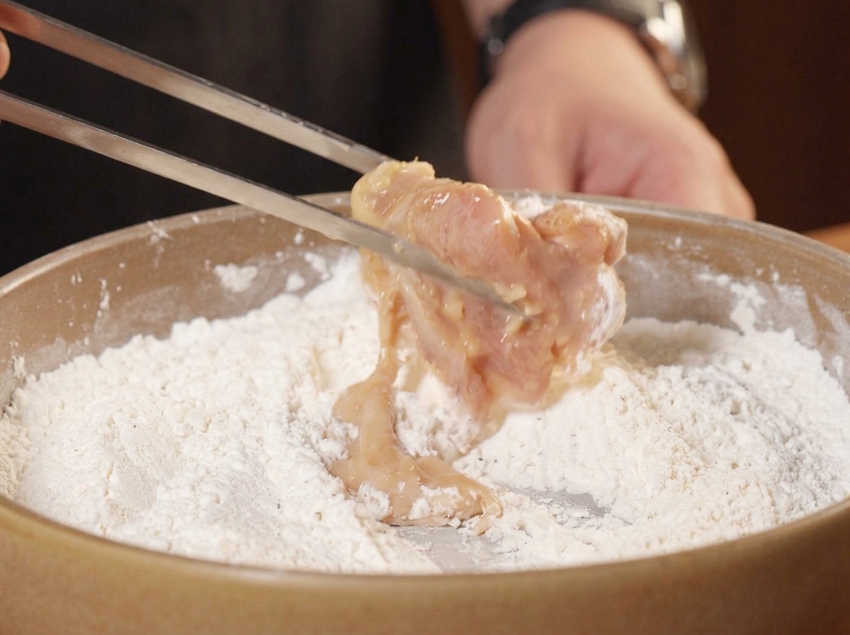 Pince déposant un morceau de poulet mariné dans le mélange de farine assaisonné.