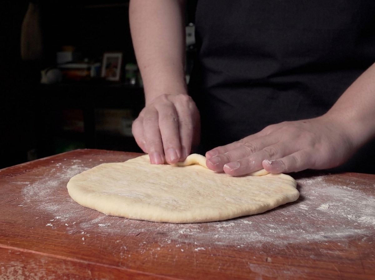 Des mains commençant à rouler le bord dune feuille de pâte plate pour former un cylindre sur un comptoir en bois.