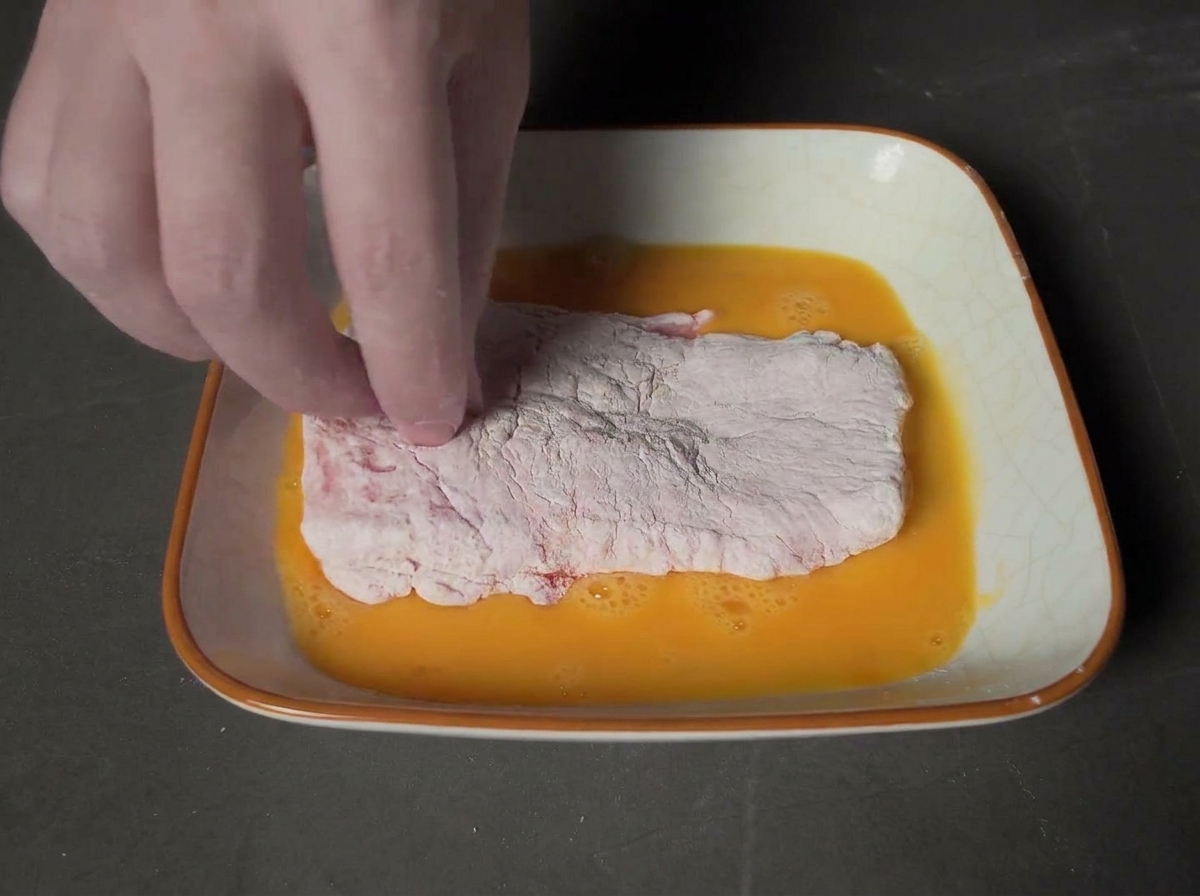 Une main trempant une côtelette de porc farinée dans un plat carré rempli dœuf battu doré.