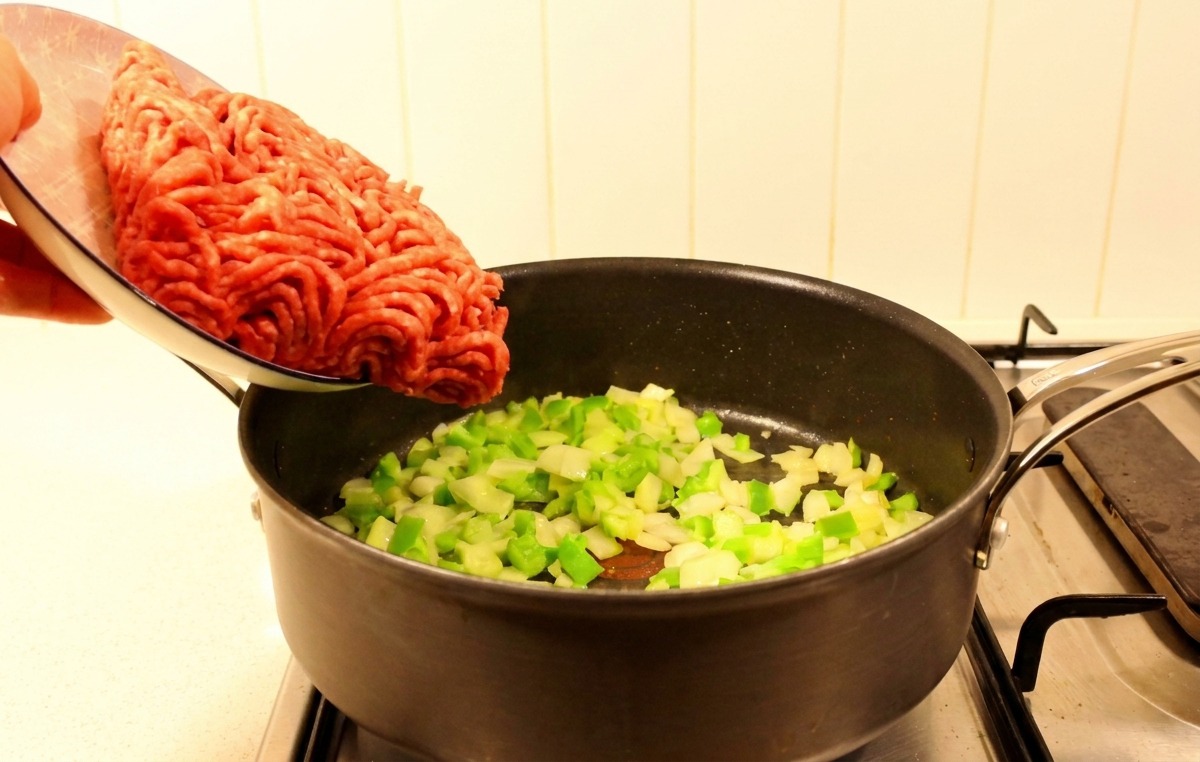 Du bœuf haché frais versé dune assiette dans une poêle contenant des poivrons et des oignons sautés.