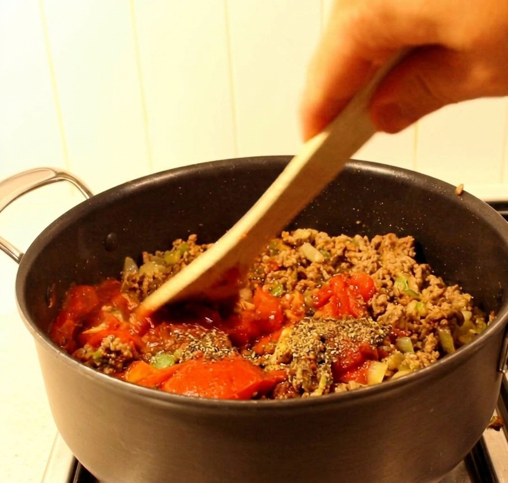 Une spatule en bois mélangeant la viande, les poivrons et la sauce tomate rouge dans une sauteuse noire.