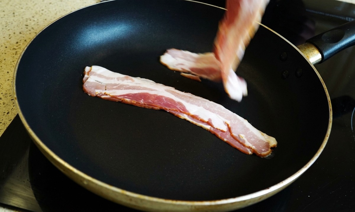Une main plaçant des tranches de bacon crues dans une poêle noire.