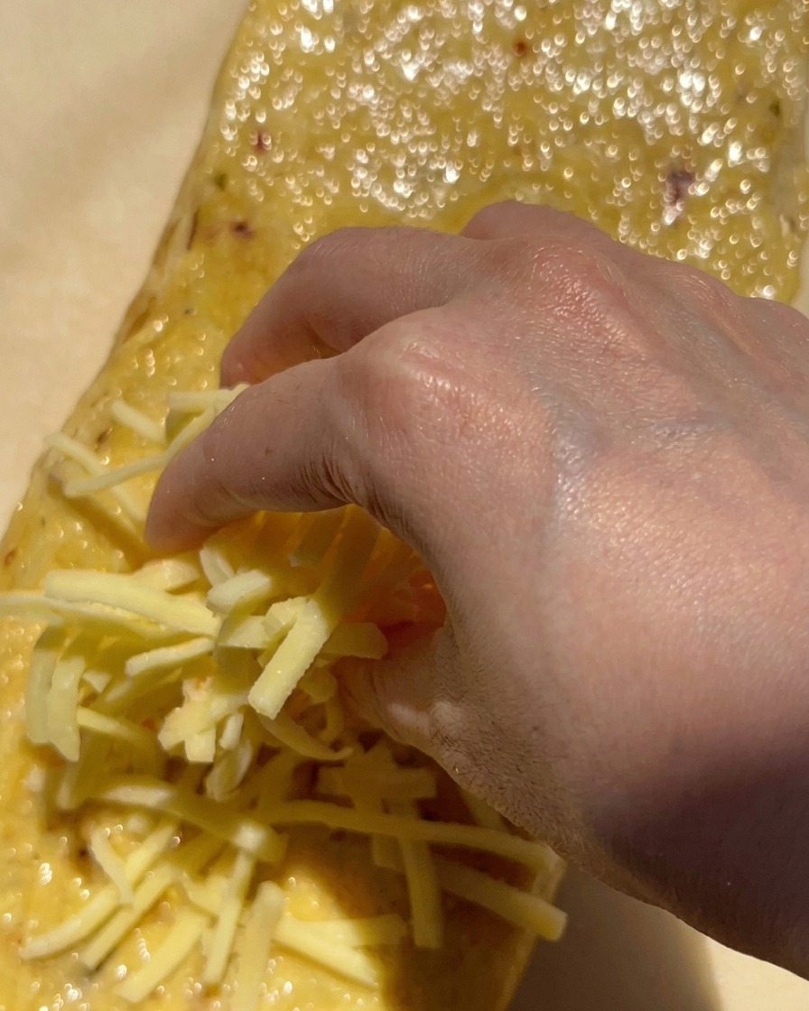 Une main parsemant une grosse poignée de mozzarella blanche râpée sur un pain recouvert de beurre dail.