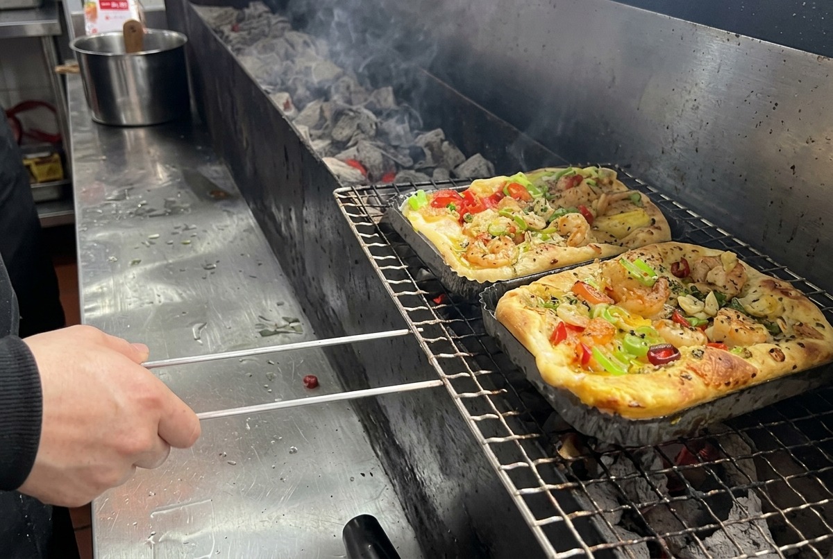 Une pizza rectangulaire garnie de crevettes et de légumes mélangés est placée sur la grille dun grand barbecue au charbon de bois dans une cuisine professionnelle.