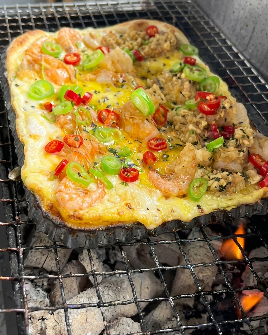Une pâte à pizza rectangulaire surmontée de crevettes et de légumes cuit au-dessus de braises de charbon ardentes.
