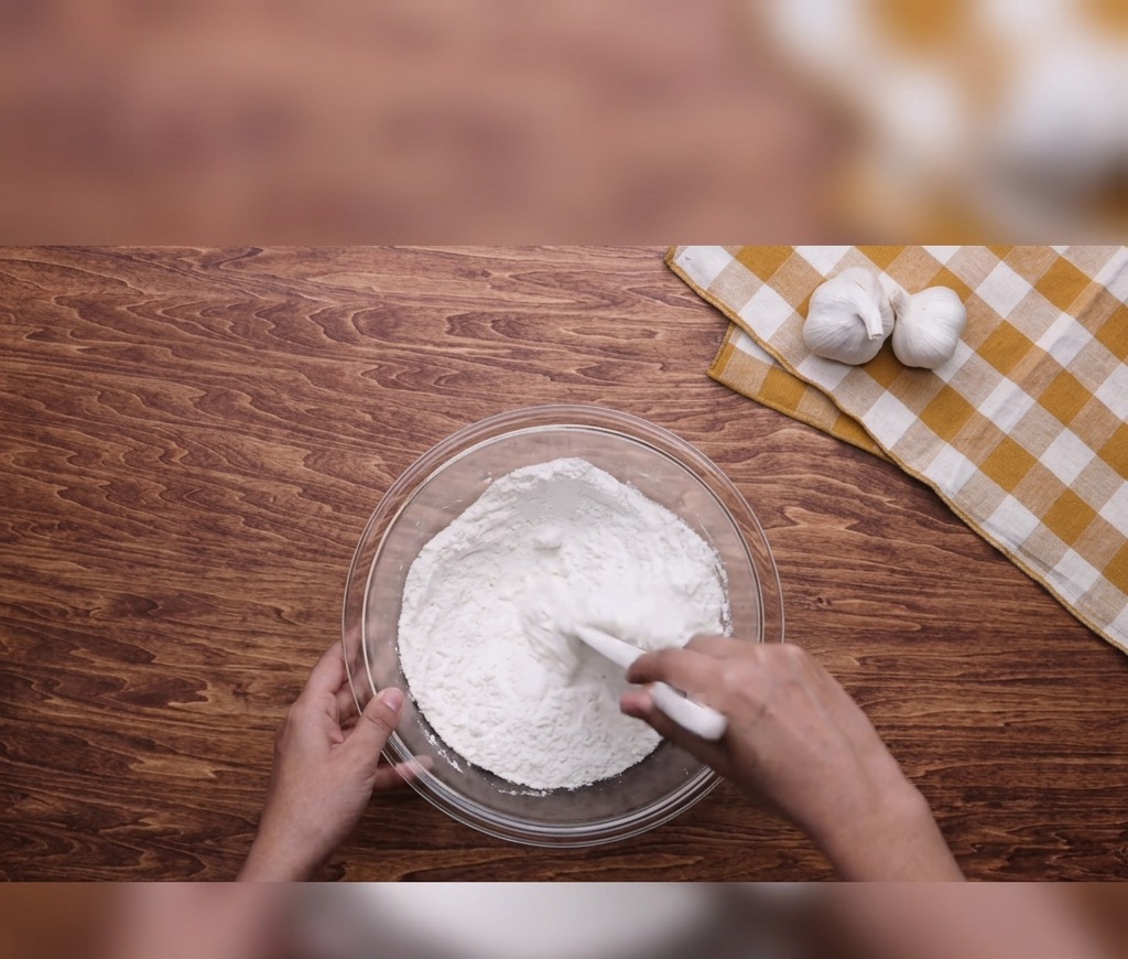 Des mains utilisant une spatule blanche pour mélanger la farine, leau et lhuile dans un saladier en verre posé sur une table en bois.