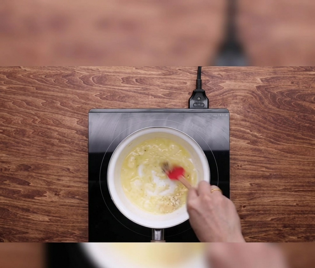 Une main utilisant une spatule rouge pour mélanger du beurre fondu et de lail émincé dans une petite casserole blanche sur une plaque à induction noire.