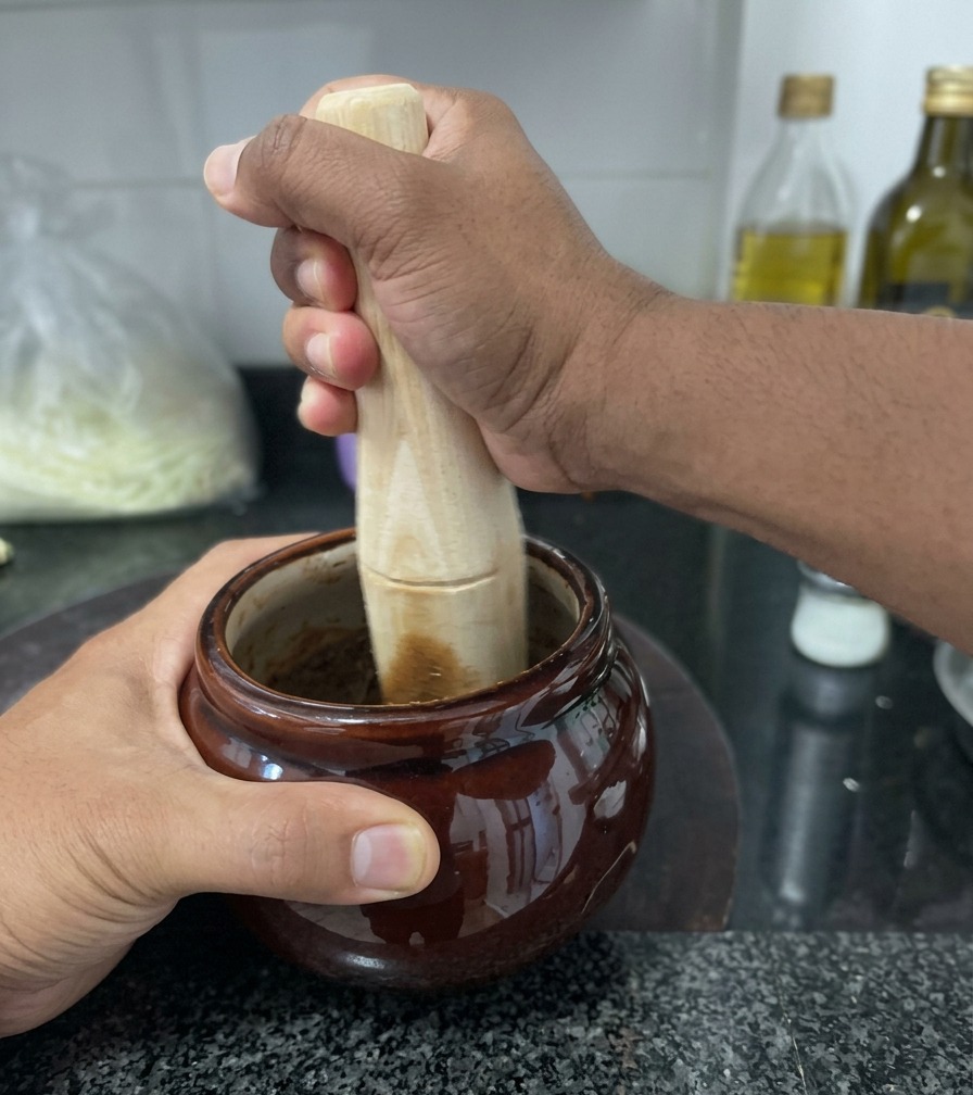 Une personne utilisant un pilon en bois pour broyer de lail frit très foncé dans un petit bocal en céramique marron sur un plan de travail en granit.