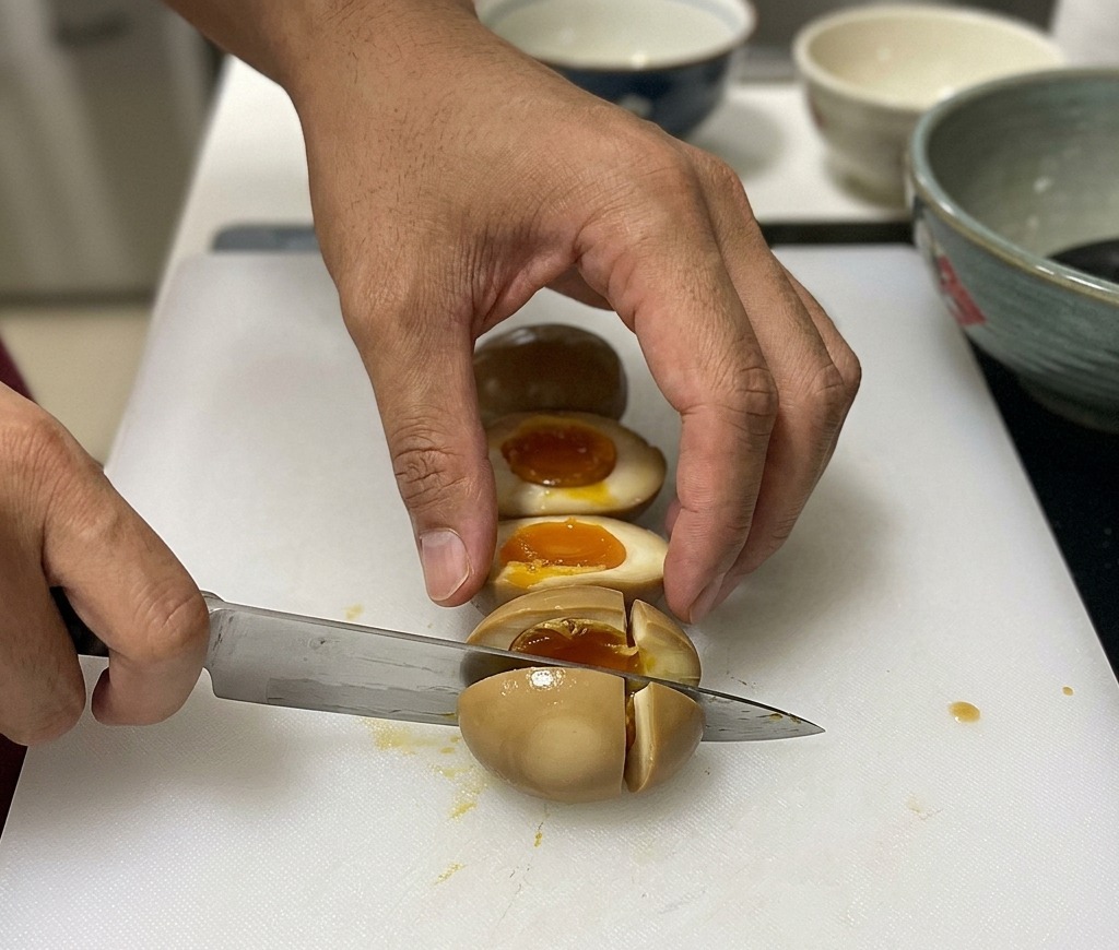Un chef coupant un œuf mollet mariné de couleur brune en deux sur une planche à découper blanche, révélant un jaune orange coulant à lintérieur.