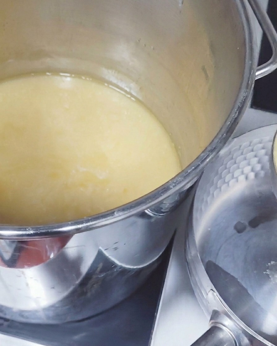 Une grande louche en métal prélevant un bouillon de porc opaque et jaune pâle dans une grande marmite pour le verser dans une casserole plus petite.