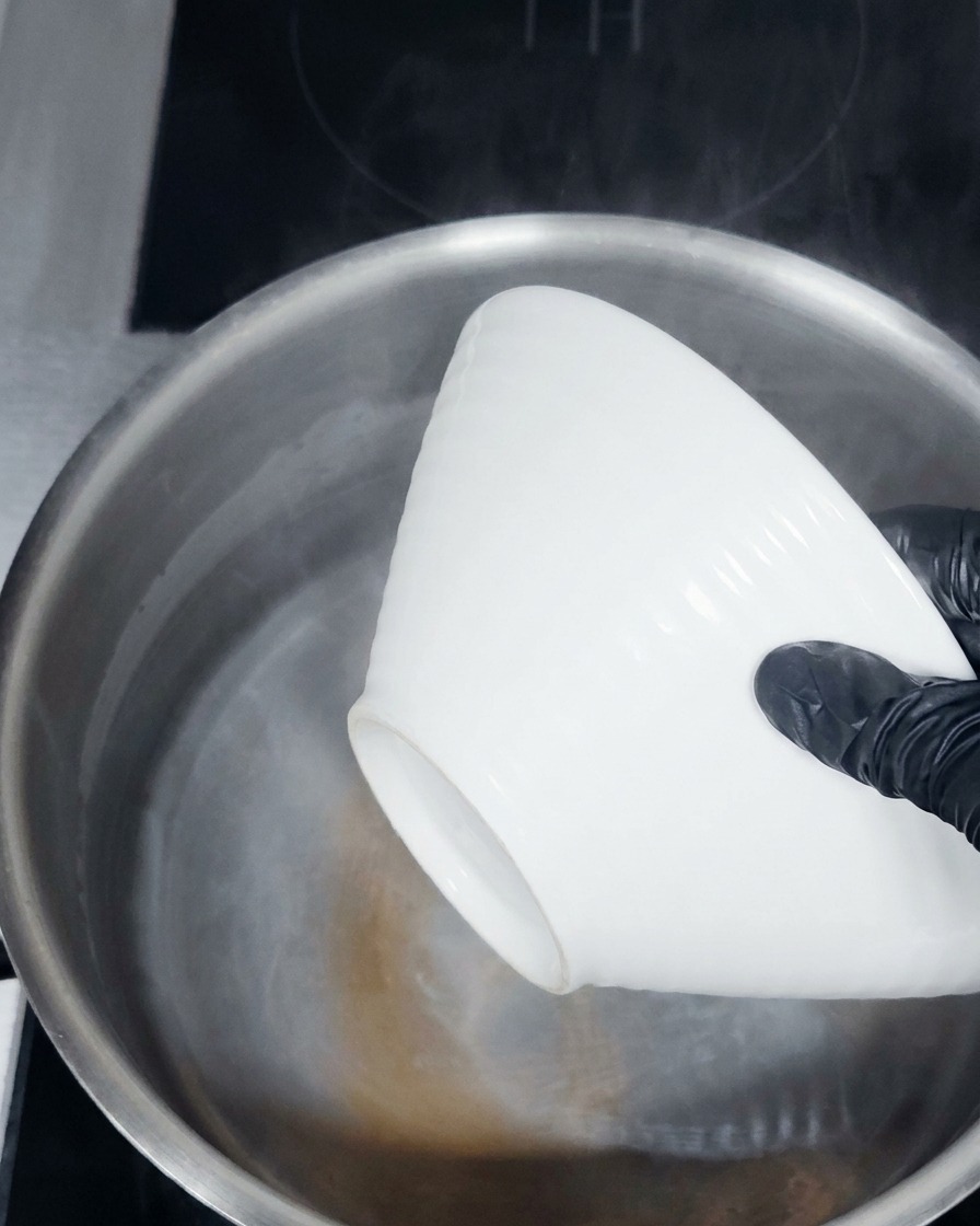 Mains gantées plongeant un bol en céramique blanche dans une casserole deau chaude fumante.
