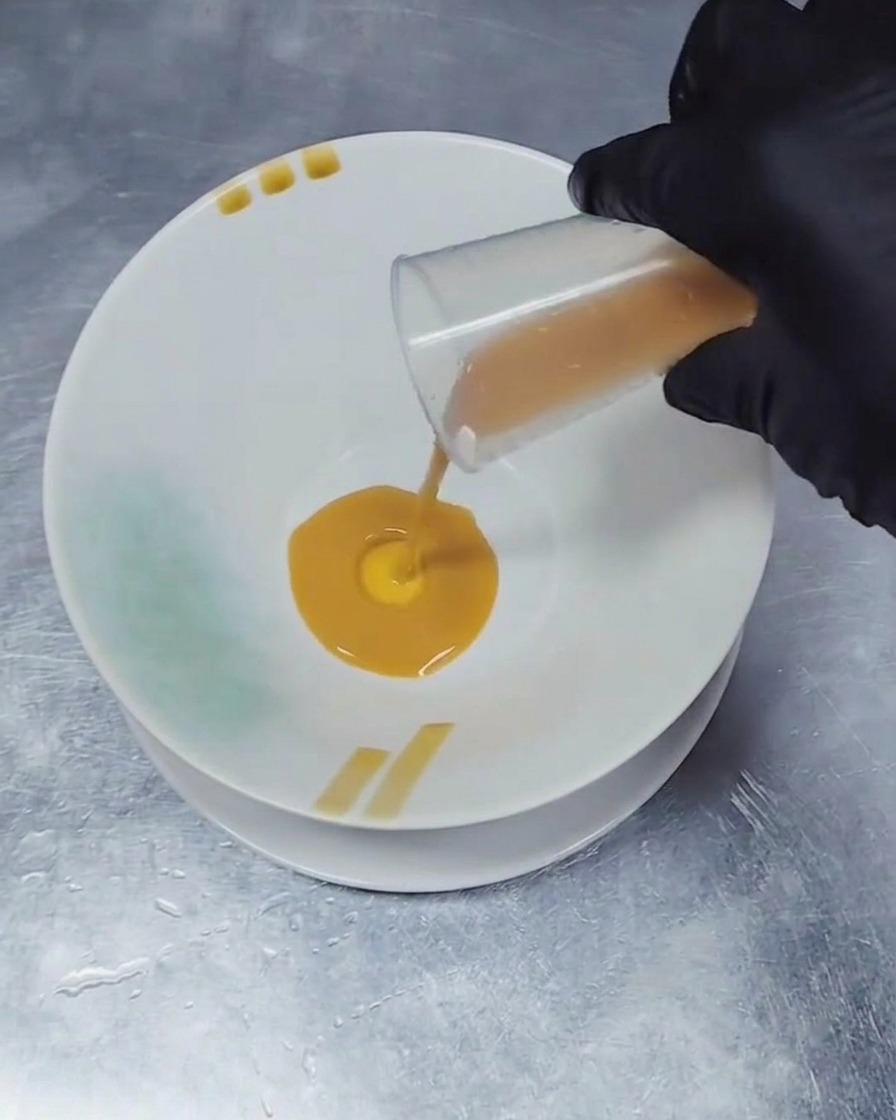 Une main gantée versant une petite quantité de base dassaisonnement liquide marron foncé depuis un bécher doseur transparent au centre dun bol en céramique blanche.