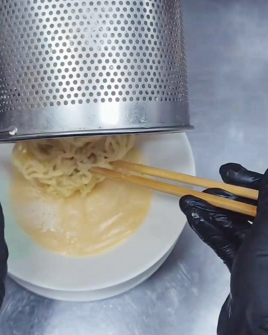 Passoire à nouilles en acier inoxydable versant des nouilles ramen ondulées dans un bol de bouillon Tonkotsu crémeux.