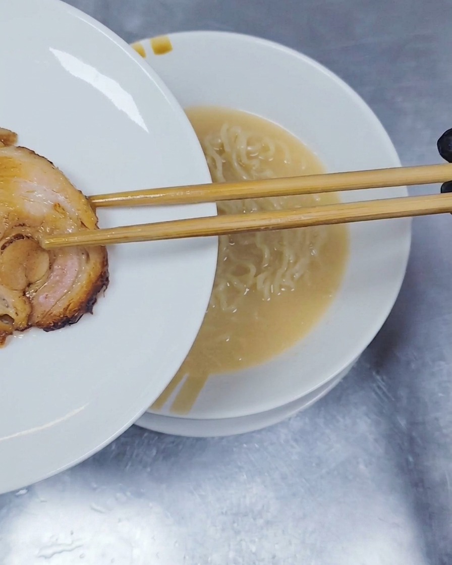 Une tranche de porc chashu roulé et braisé déposée sur les nouilles ramen à laide de baguettes en bois.