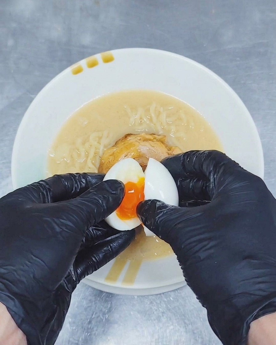 Une paire de mains gantées ouvrant un œuf mollet en deux au-dessus dun bol de ramen pour révéler un jaune doré et coulant.