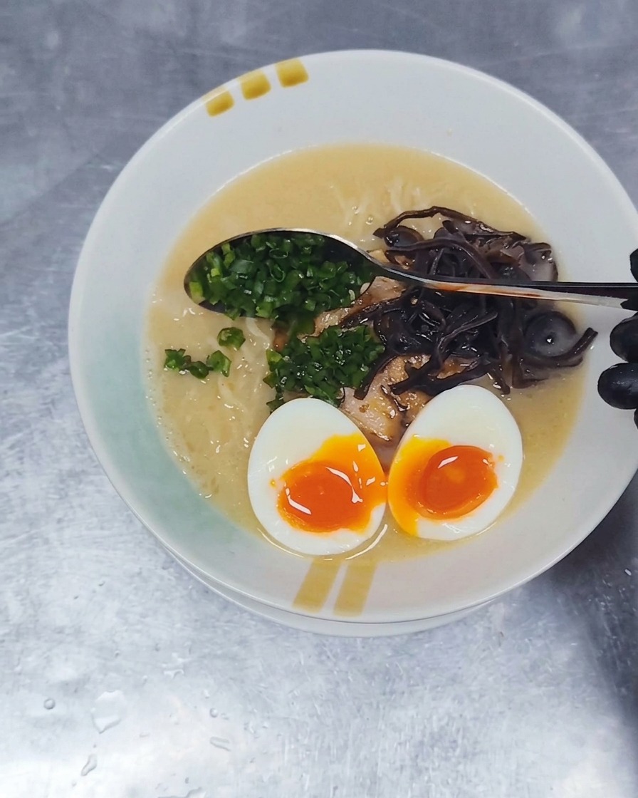 Une cuillère en métal parsemant des oignons verts finement ciselés à la surface dun bol de ramen Tonkotsu.