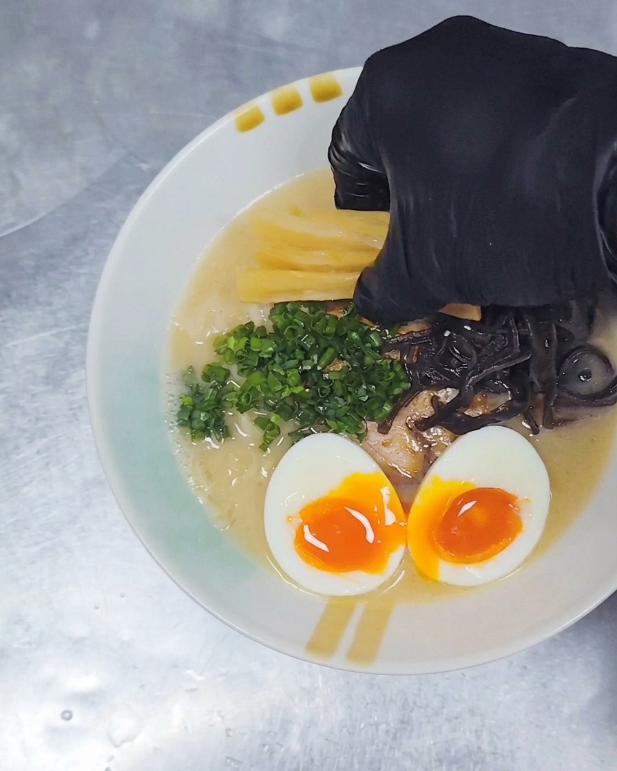 Une main gantée déposant des lanières jaunes de pousses de bambou assaisonnées sur un bol de ramen déjà garni de porc, dœuf et doignons verts.