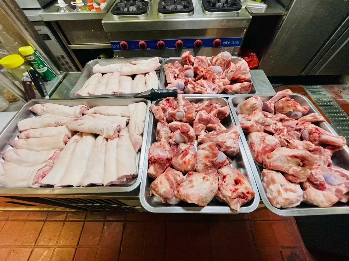 Quatre plateaux métalliques remplis de jarrets et de pieds de porc crus disposés sur un plan de travail en acier inoxydable.