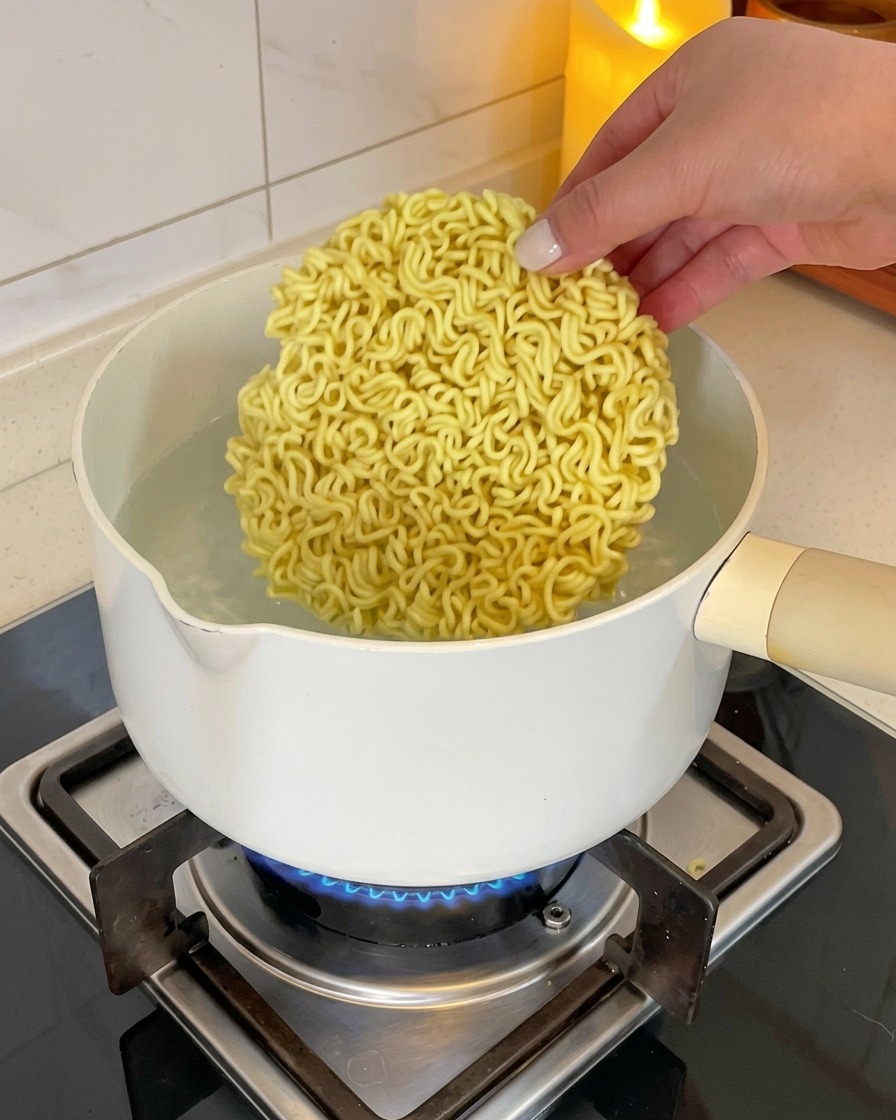 Une main dépose un bloc circulaire de nouilles ramen instantanées dans une casserole deau bouillante.