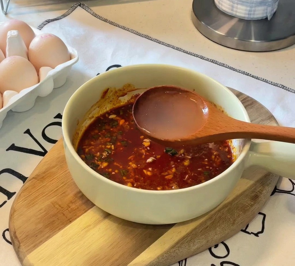 Une louche en bois verse de leau de cuisson chaude dans un bol contenant une pâte dassaisonnement orange foncé.