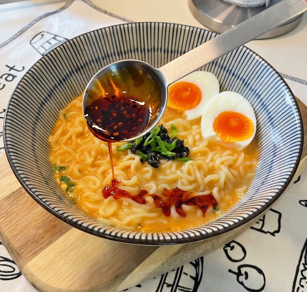 Une cuillère doseuse en métal verse de lhuile piquante rouge vif contenant des flocons de piment visibles dans un bol de ramen préparé.