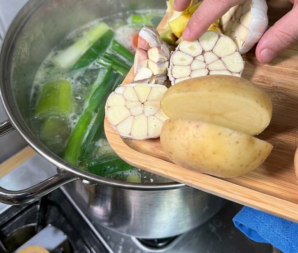 Une main ajoutant des têtes dail coupées en deux et des pommes de terre depuis une planche en bois dans un bouillon fumant.